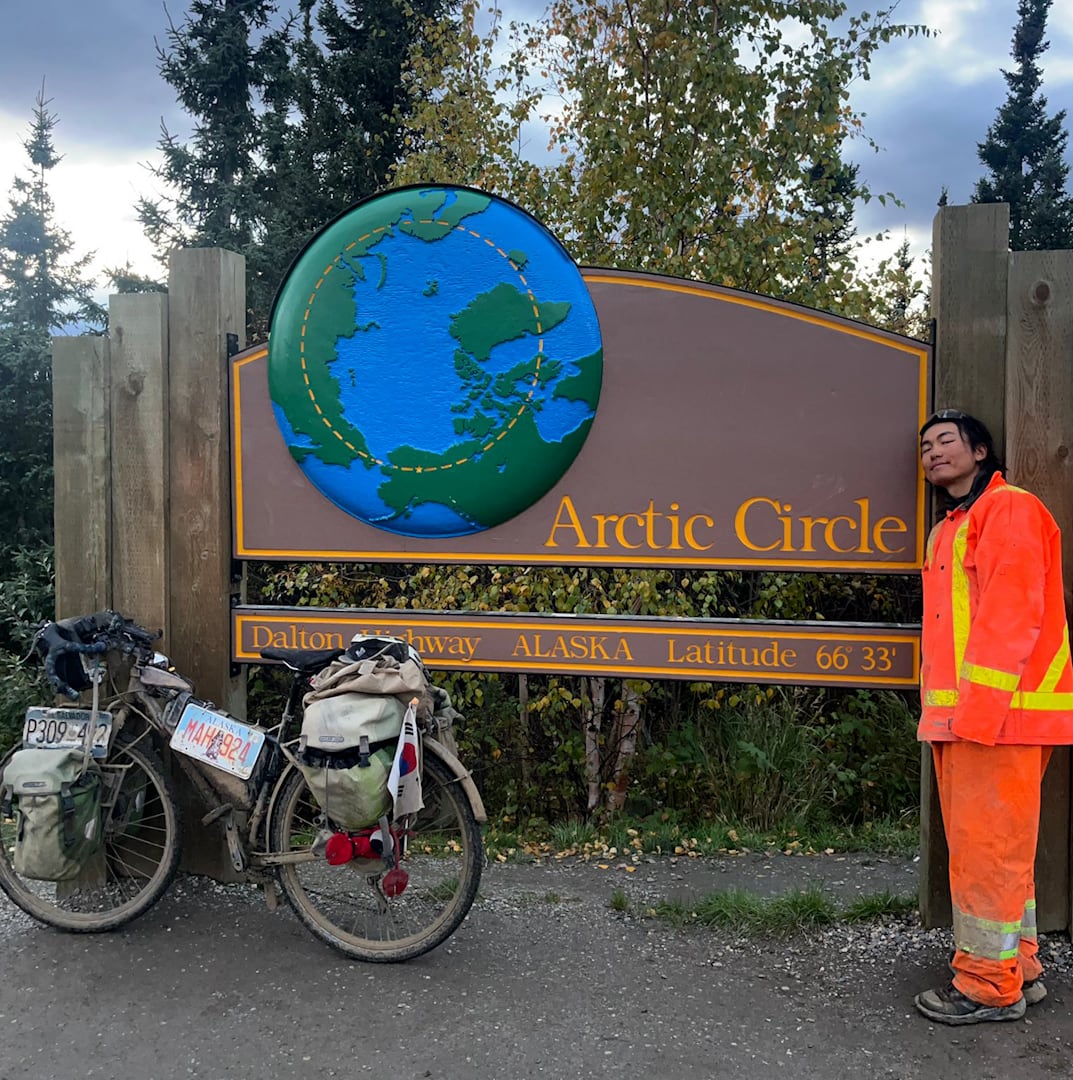 A man dressed in orange stands in front of a sign that says Alaska