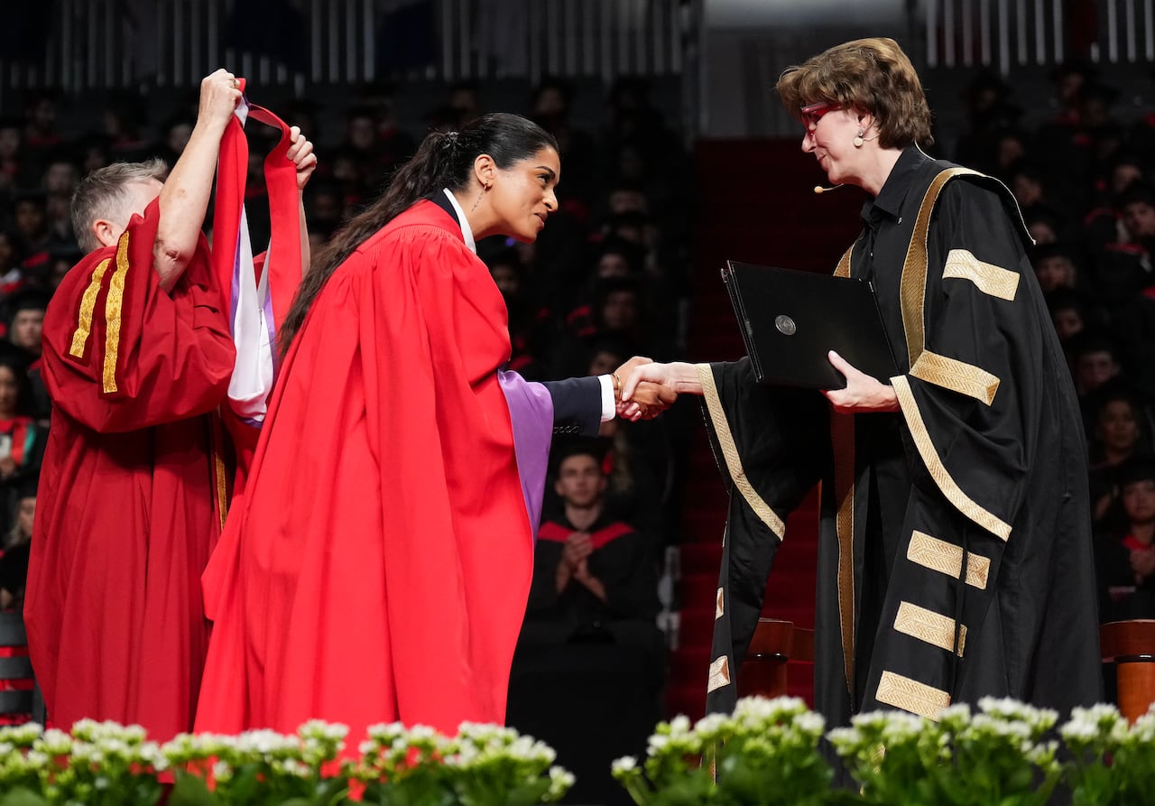 A woman in black robes shake hands with Lilly Singh on a graduation stage, while another woman goes to place a ceremonial hood on Singh.