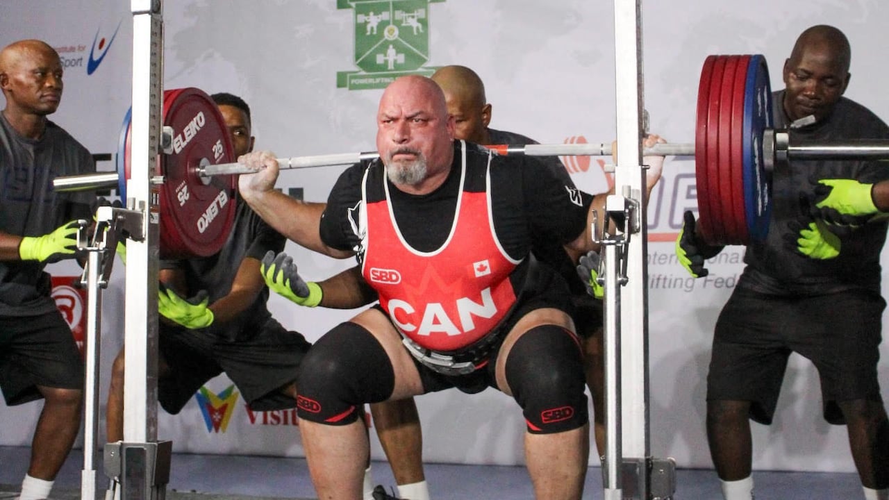A man lifts a barbell with heavy weights over his shoulders with a man on either side of him for support.