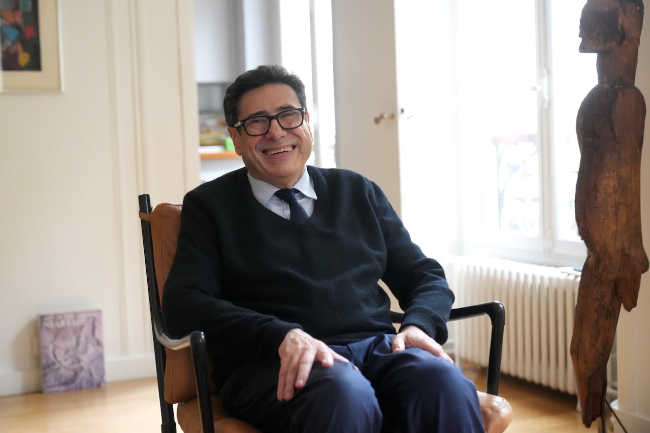 A man wearing a black V-neck sweater and glasses smiles while posing for a photo while sitting in a chair.