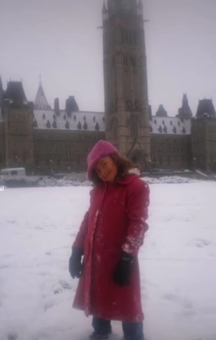 A young girl with a winter coat stands in the snow, head slightly cocked. Behind her is Parliament Hill's centre block building.