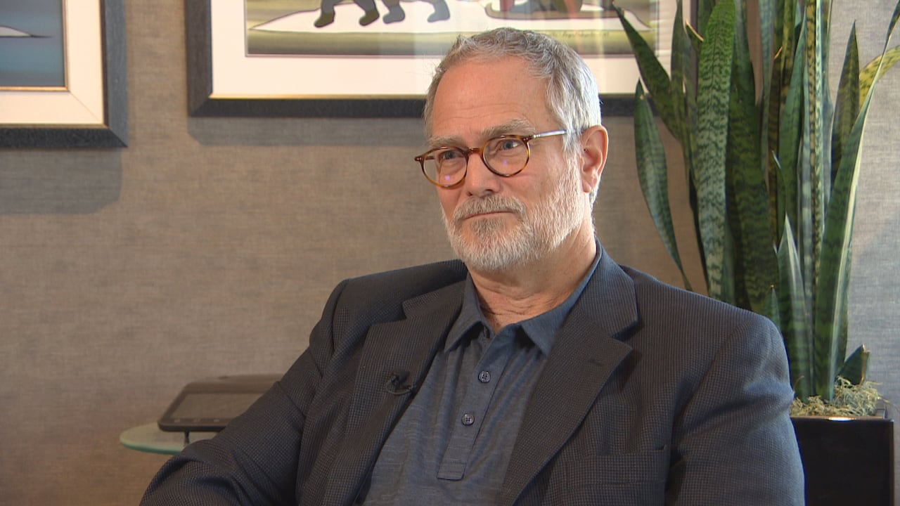 A man with glasses sits with a plant behind him.