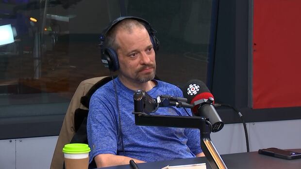 A man in a blue shirt sits behind a radio mic in a studio