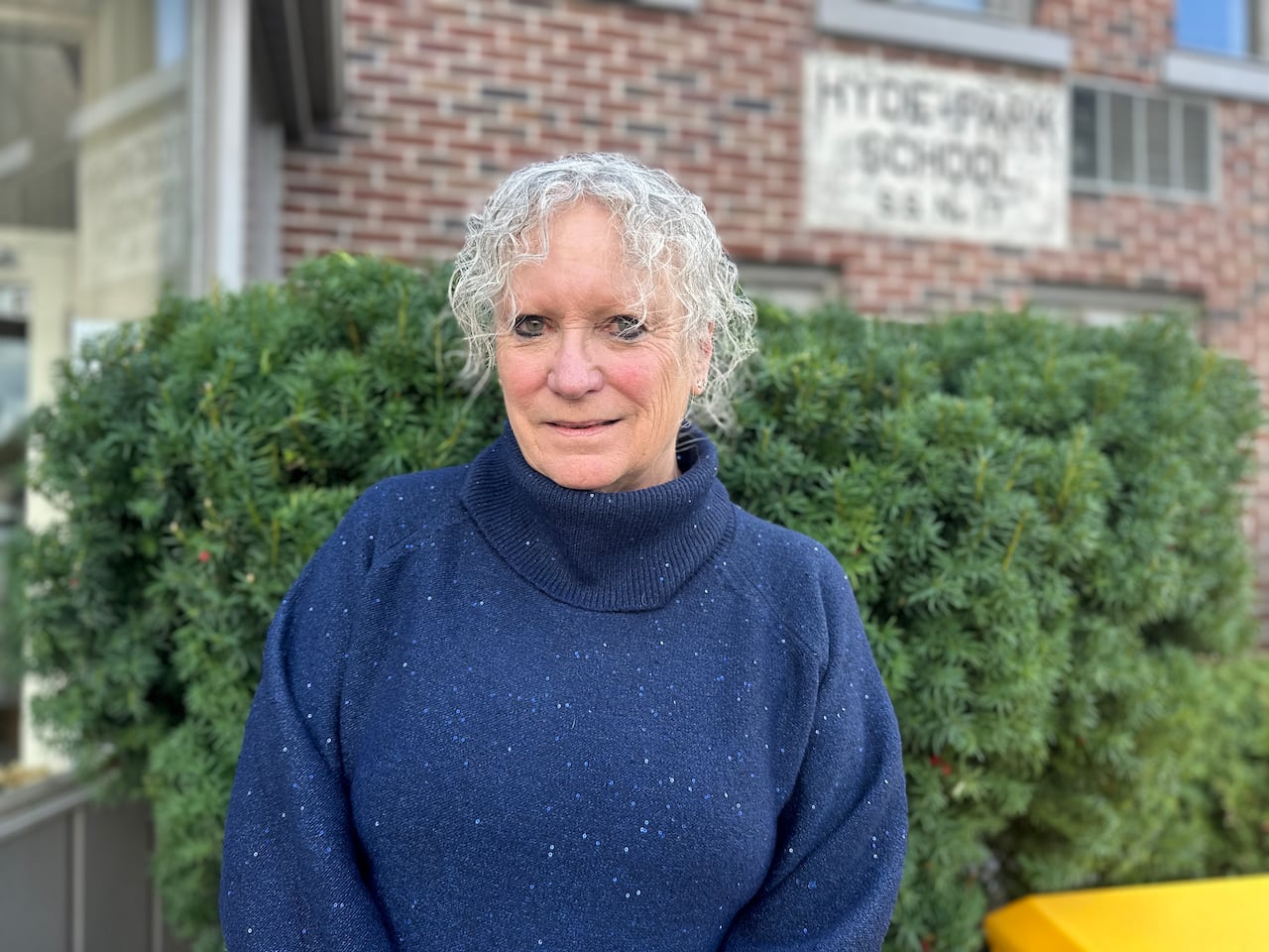 A pistillate with grey hairsbreadth smiles astatine the camera. She is wearing a bluish sweater and lasting successful beforehand of a greenish bush.