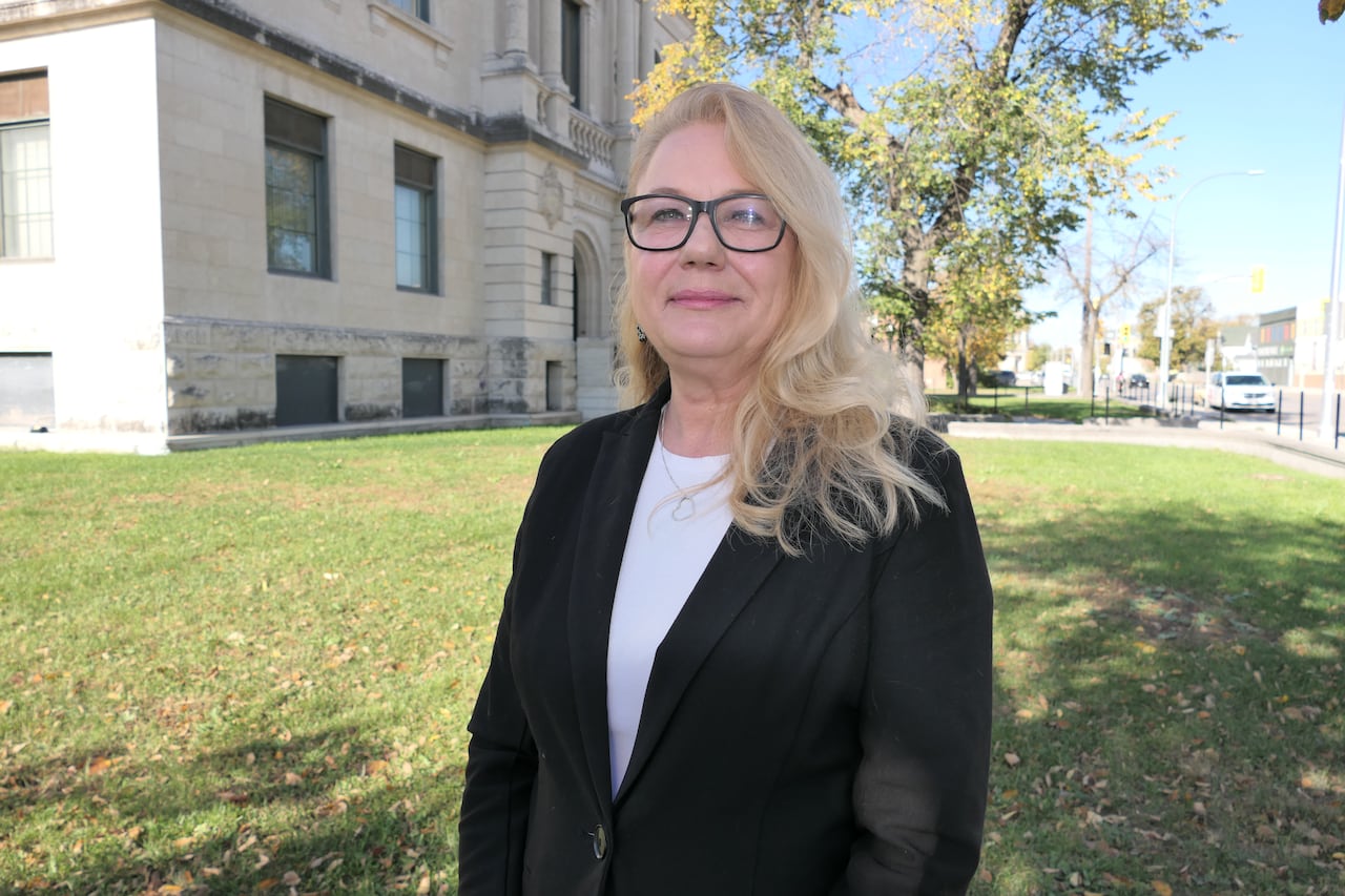 A woman with long blonde hair, black eyeglasses and a black blazer stands outside
