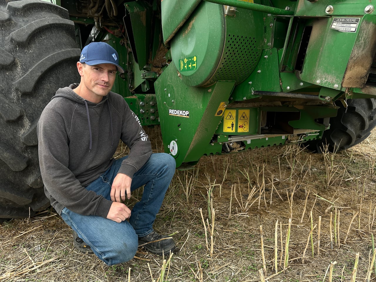 A man in a blue hat, grey hoodie and blue jeans kneels next to a green combine with a Redekop brand add on.