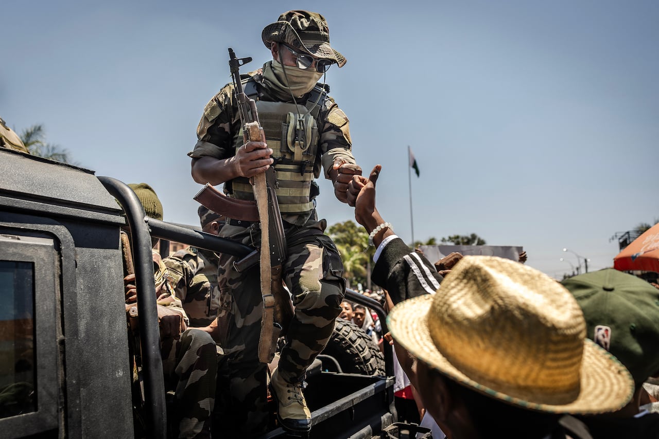 An armed soldier on the back of a pickup truck rams a civilian on the road while a crowd gathers around him.