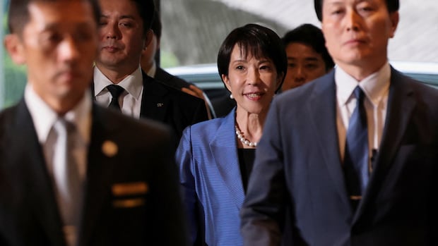 Sanae Takaichi arrives at the prime minister's official residence in Tokyo on Tuesday. A woman in a blue suit, wearing pearl, walks surrounded by men in dark suits.