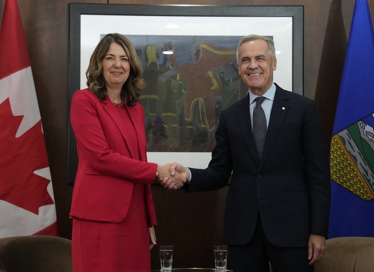 A woman in a red suit and a man in a black suit shake hands in front of a Canadian and Albertan flag.