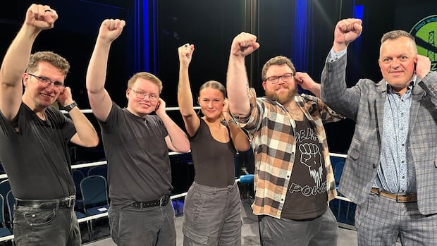 Wrestler Justin Marriott a.k.a Deaf Power, second from right, wants the sport to be more inclusive. He poses with several interpreters and the owner of Kaizer Pro Wrestling. Five people stand together in a wrestling ring and each hold one fist to their ear and one to the sky.