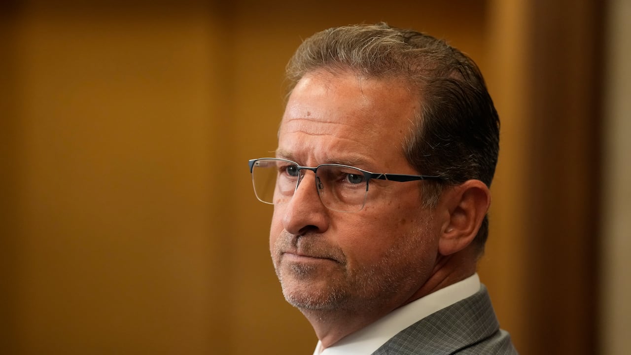 A man in a grey suit and glasses speaks in a foyer.