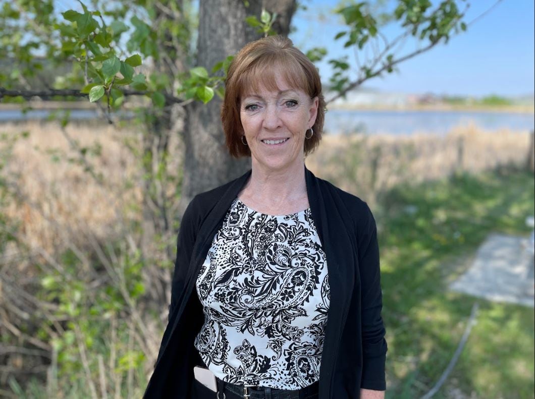 A pistillate outdoors successful beforehand of a histrion and grassy area, smiling astatine the camera successful a achromatic overgarment and patterned shirt.