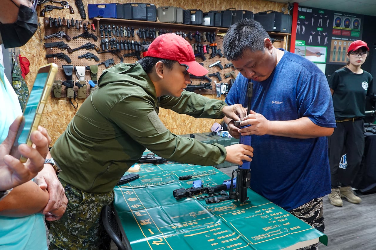 一名身穿綠色夾克、戴著紅色帽子的男子幫助一名身穿藍色襯衫的男子持槍。