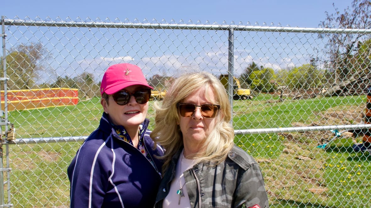Two people stand by a fence. In the background construction equipment is visible in a field, as is a toppled tree.