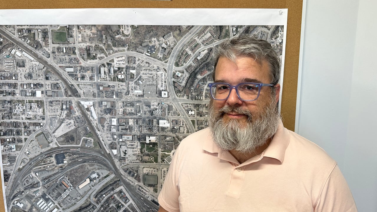 A antheral   with a chaotic  grey beard and hairsbreadth  wearing a pinkish  garment  stands successful  beforehand   of a representation  of downtown Sudbury