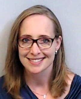 A woman with shoulder length brown hair and glasses is earing a navy blouse and smiling