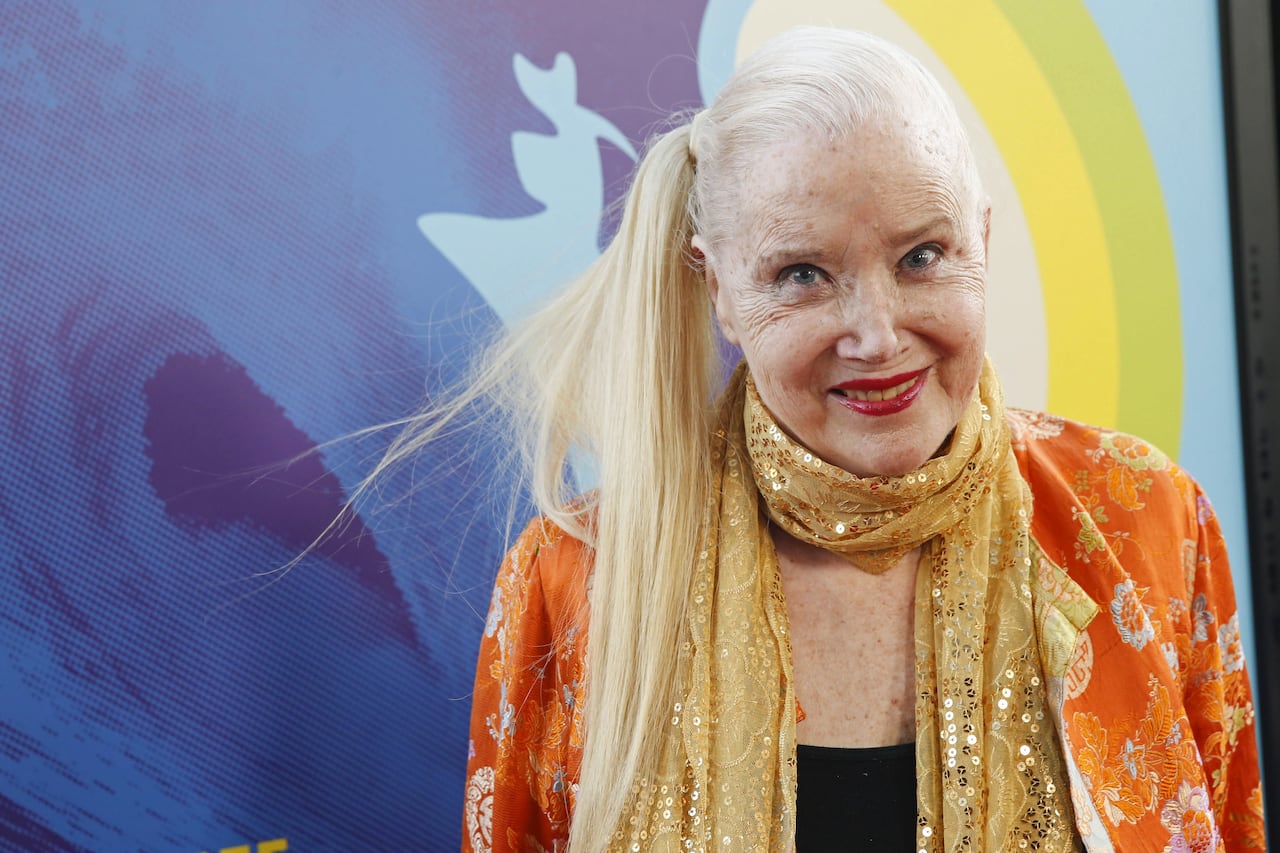 An elderly woman with blonde hair in a long ponytail and a beaded scarf smiling at the camera
