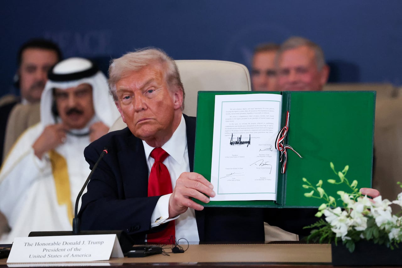 A man wearing a dark suit and red tie holds up a green folder with a signed white document inside. He is seated at a table with a small microphone in front of him, with others gathered behind.