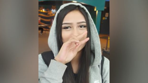 A woman with brown hear wearing a hoodie. She's holding two fingers up with her right hand, which is covering parts of her face.