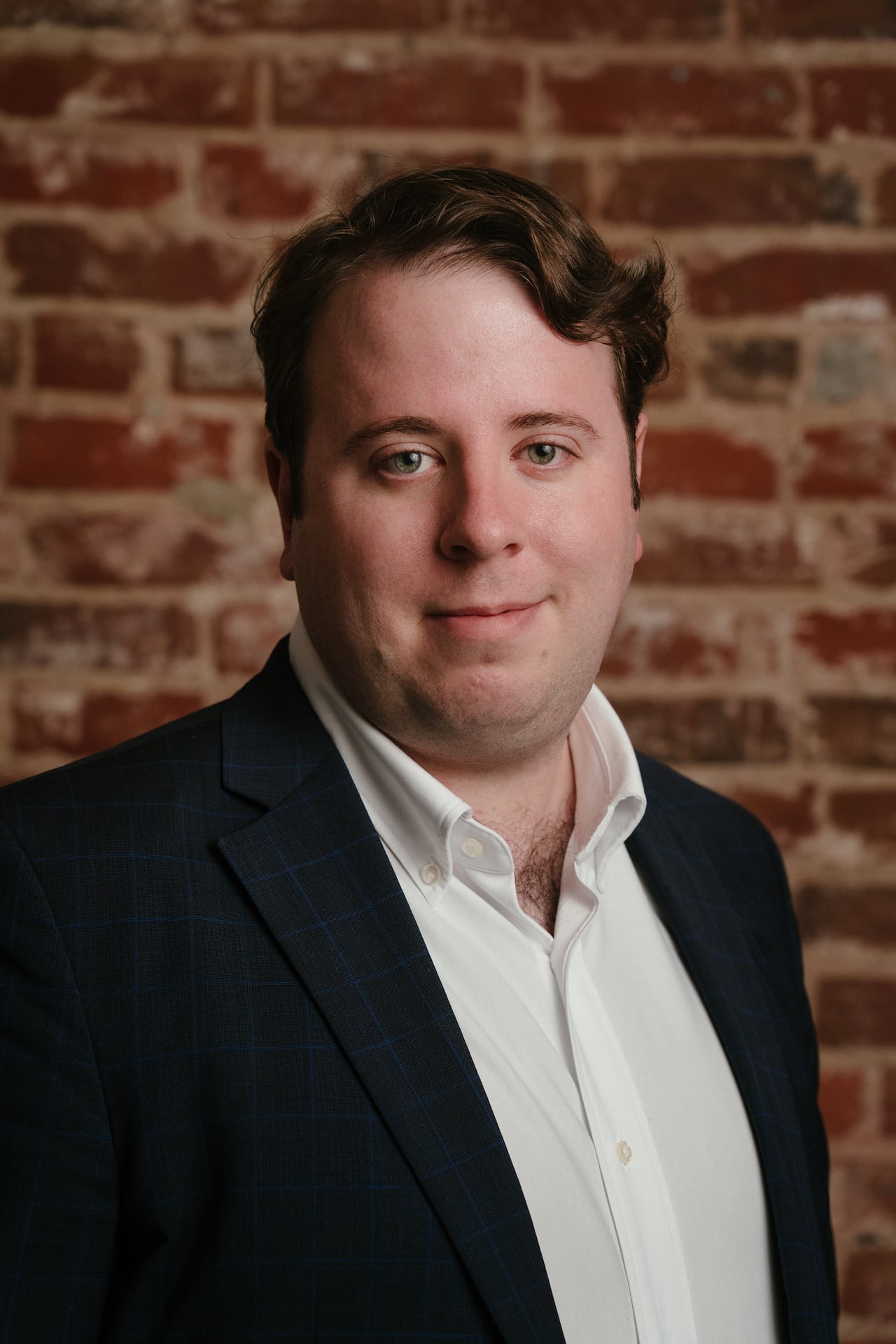 A man in a black blazer and white shirt stands in front of a brick wall looking at the camera.