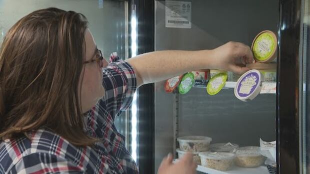 Someone puts cheese on a fridge shelf in a store. A woman in a plaid shirt and glasses puts round containers of cheese on a fridge shelf in a store.