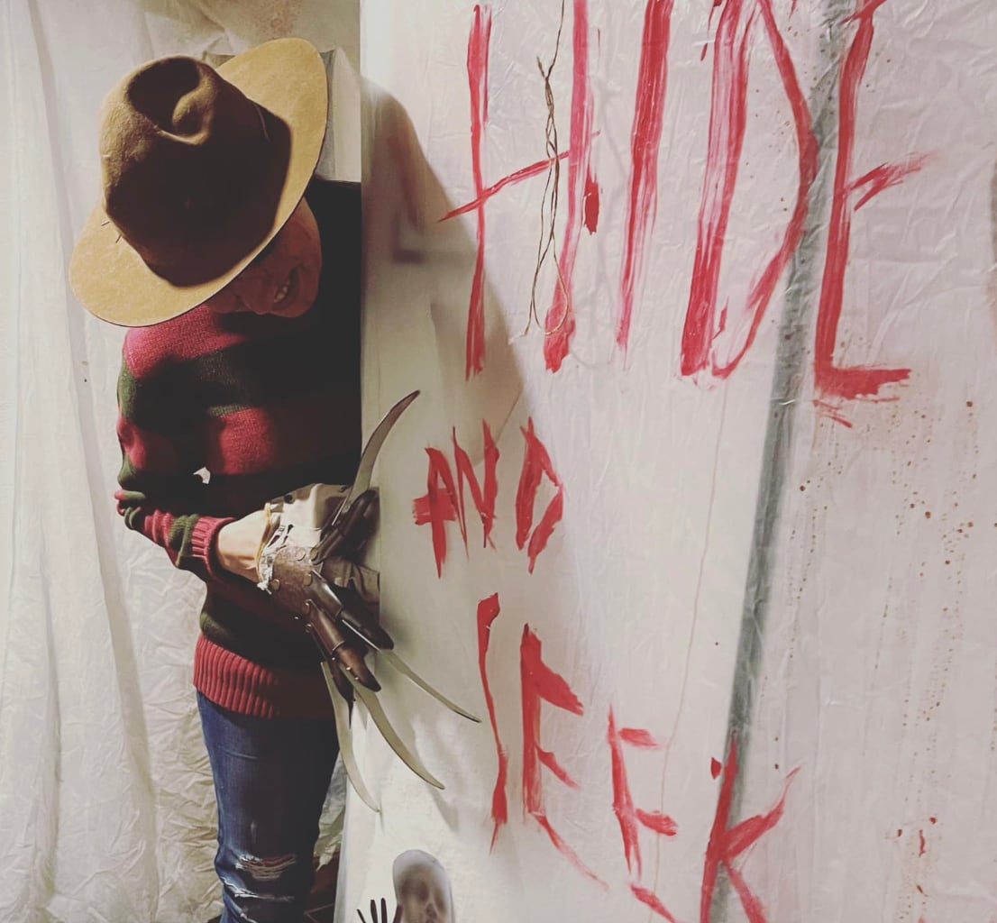 A man dressed as Freddy Krueger, wearing a brown hat, a red and green striped sweater and a glove with knives for fingers, stands next to a white sheet with "Hide and seek" written on it in fake blood.