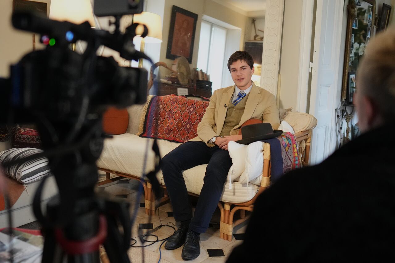 A man in a suit and fedora sits on a couch and speak to cameras