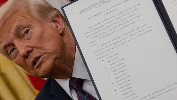 U.S. President Donald Trump looks on as he issues executive orders and pardons for Jan. 6 defendants, whose names are seen on the document he holds, in the Oval Office at the White House on Inauguration Day in Washington on Jan. 20, 2025. A man in a suit holds up a document
