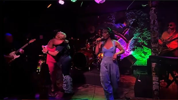 Woman in blue singing while two other women hug on stage