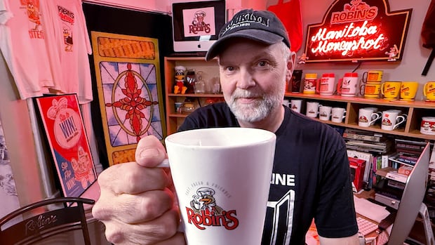 Man holds coffee cup toward camera while stand in front of Robin's Donuts memorabilia 