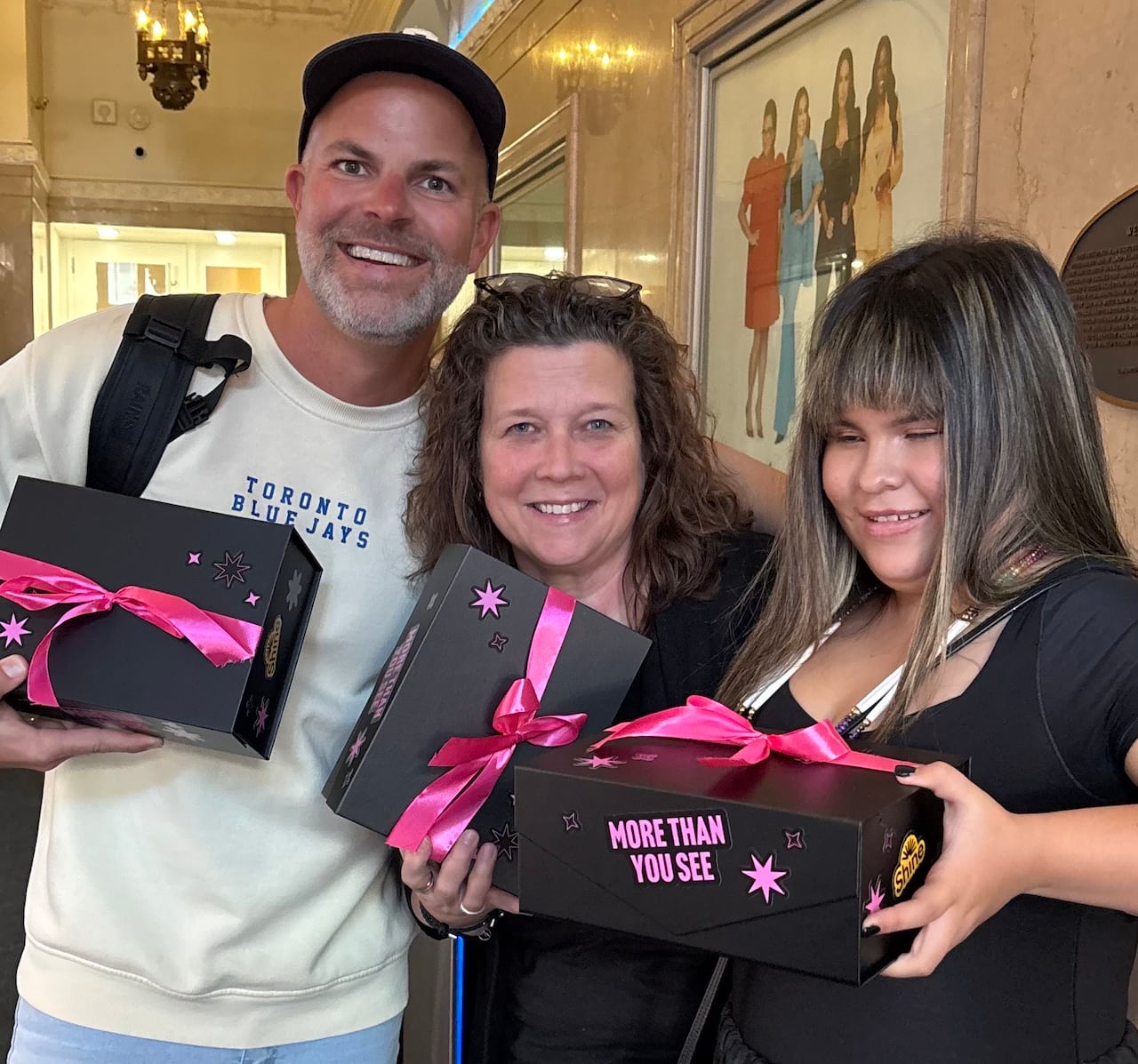 Three people stand smiling at the camera holding black boxes with pink bows tied around them
