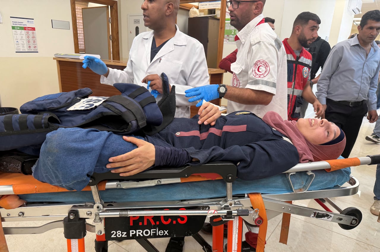A Palestinian woman, wearing a dark blue dress and a pink headscarf, puts a blue flak jacket over her waist as two men in white uniforms stand behind her in the hospital.