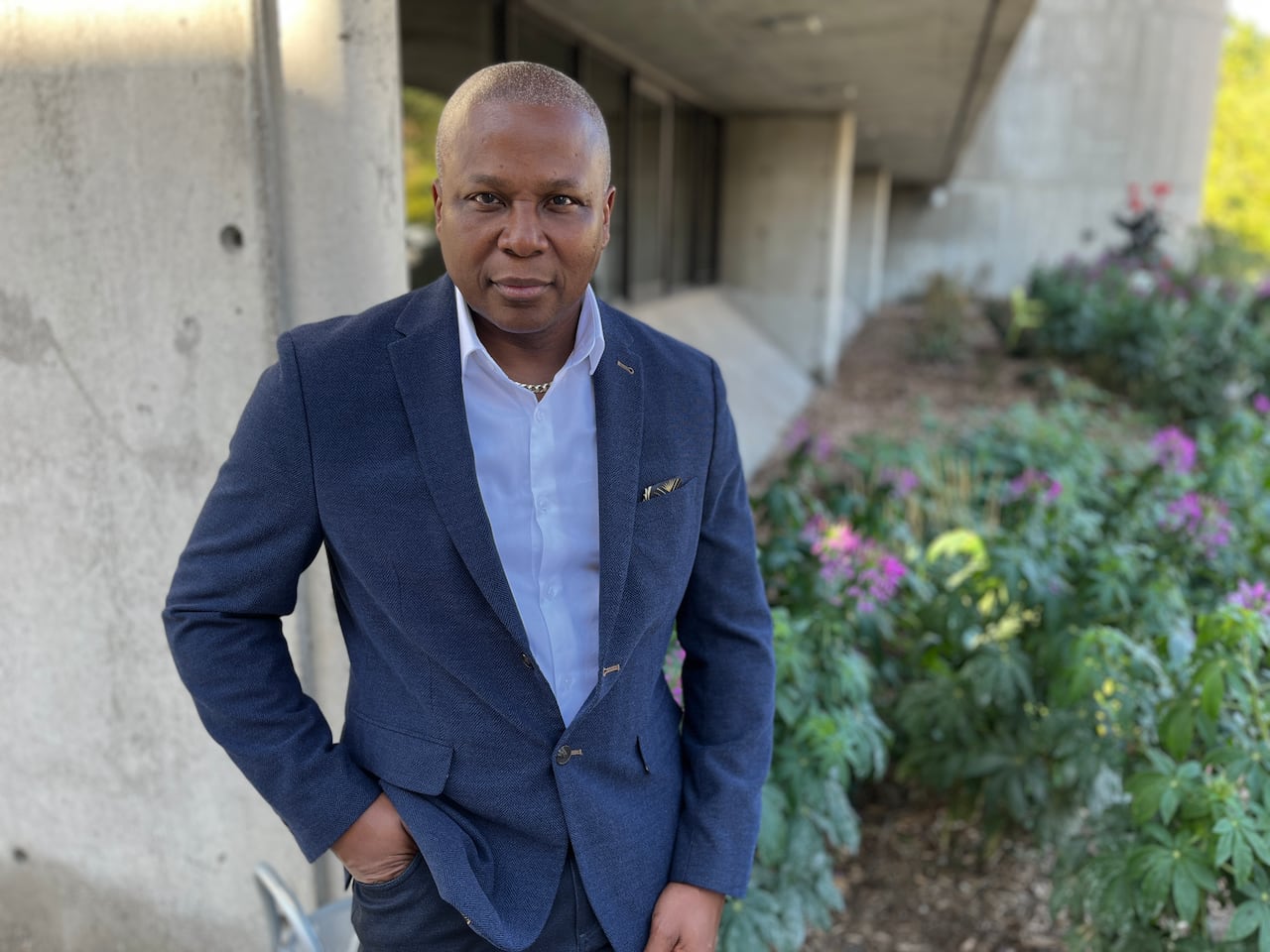A black man with very short hair is in a blue blazer and white shirt stainding in front of a concrete buidling and small bushes.