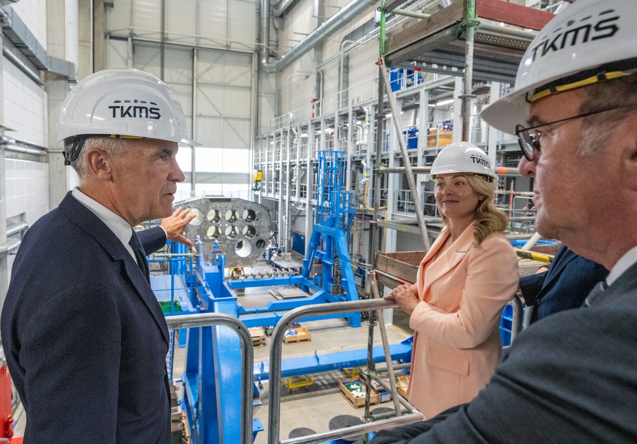Two men and a woman wearing helmets point to factory equipment.