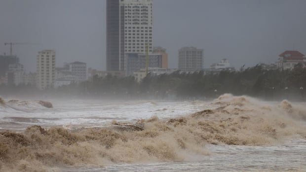 Raging brown waves crash against a city shoreline, with tall buildings in the background under a dark, stormy sky. The scene conveys intense turmoil.