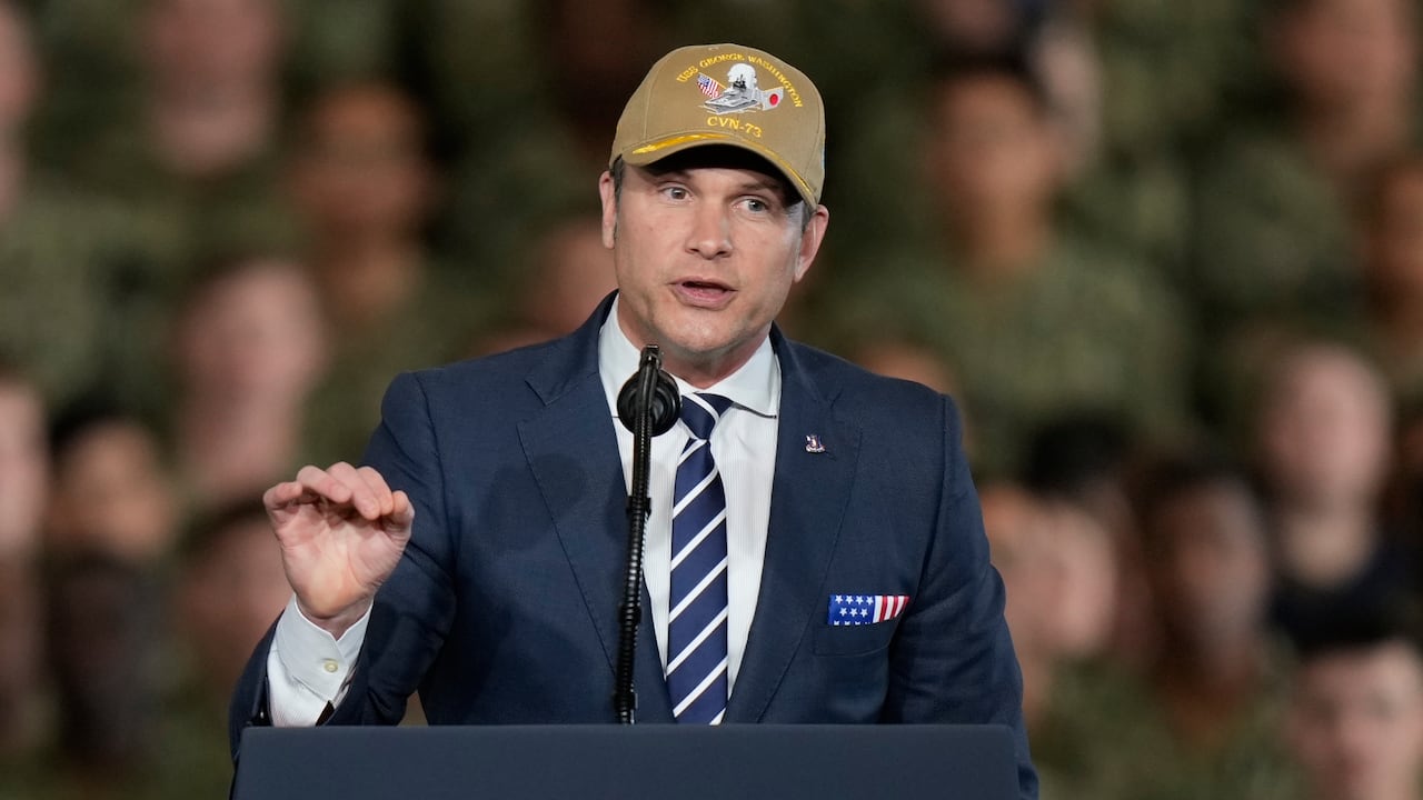 A cleanshaven man wearing a baseball cap and suit and tie is shown gesturing and speaking behind a podium.
