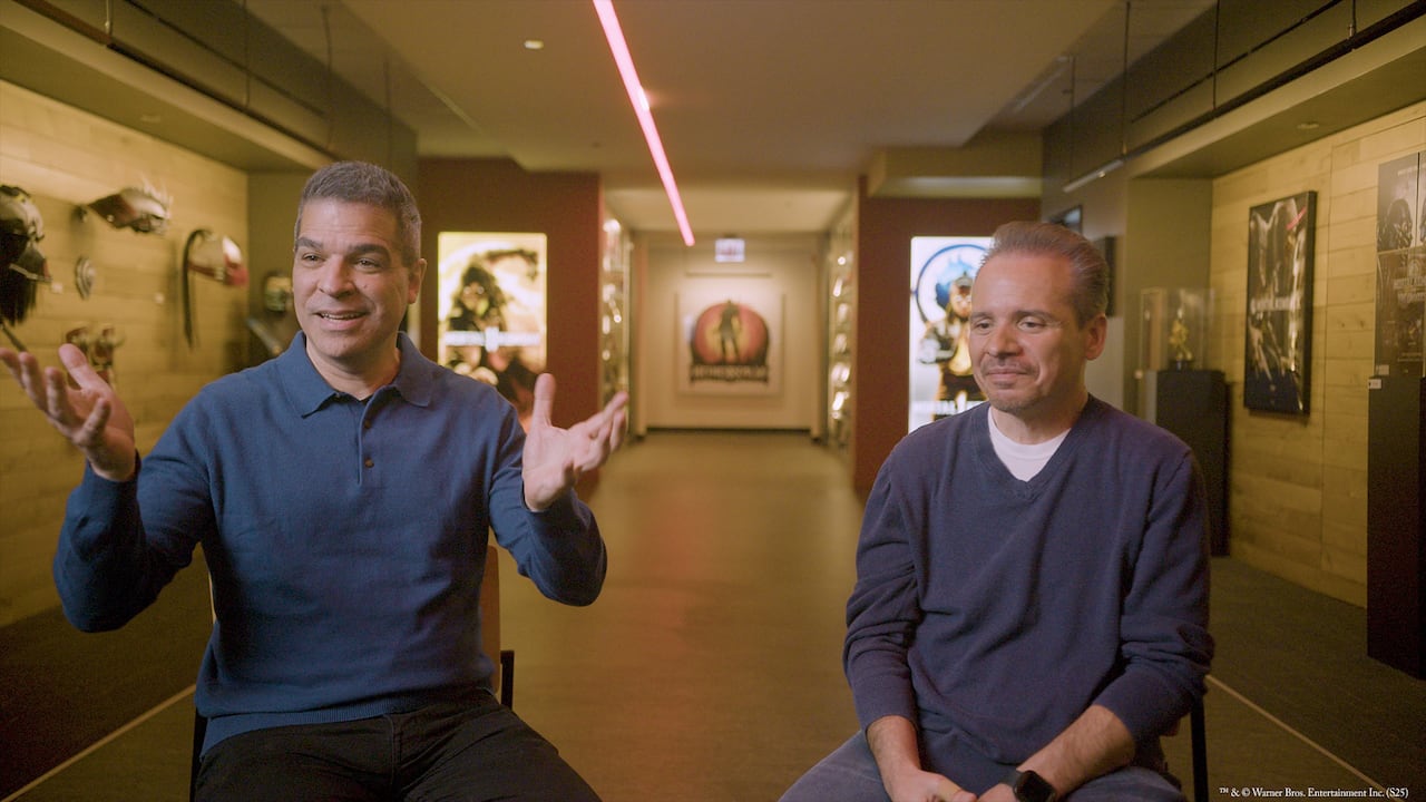 Two adult men with greying hair sit on chairs in a studio. Art and paintings are seen on the background walls.