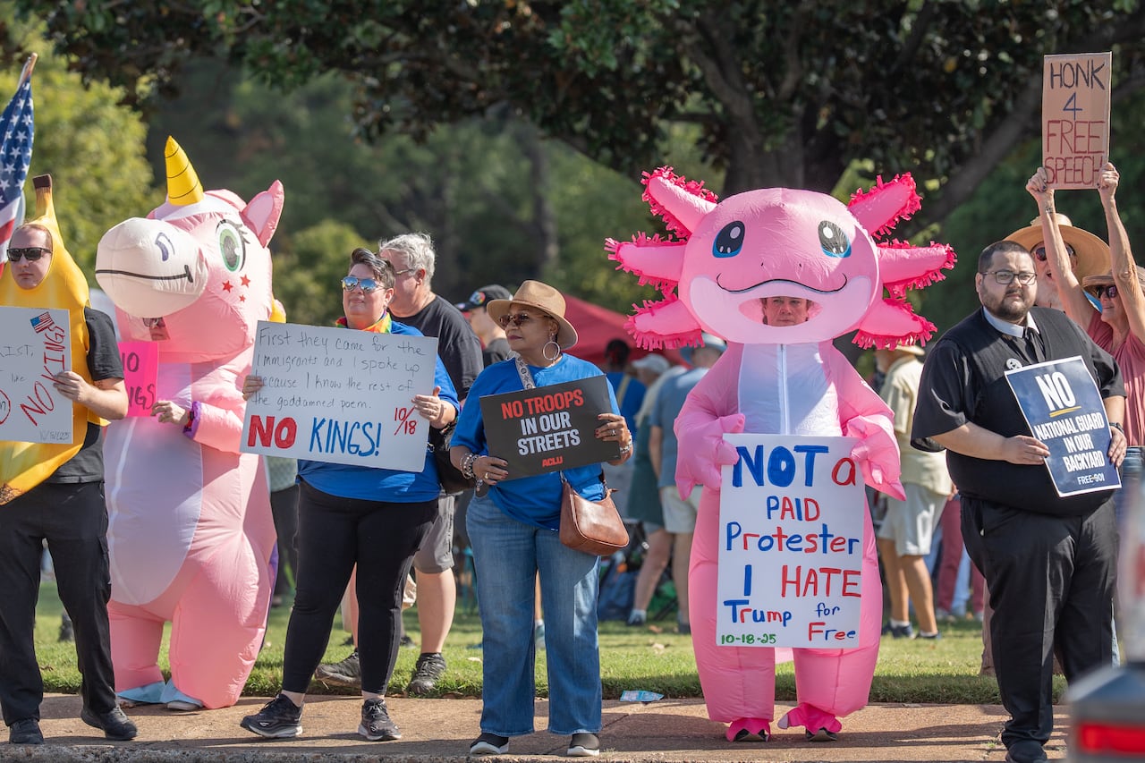 Protesters stand side-by-side. Among them, a person in a pink inflatable axolotl costume with a sign that reads" "Not a paid protester. I hate Trump for free," a person in a pink inflatable unicorn costume, and a man dressed as a banana. 