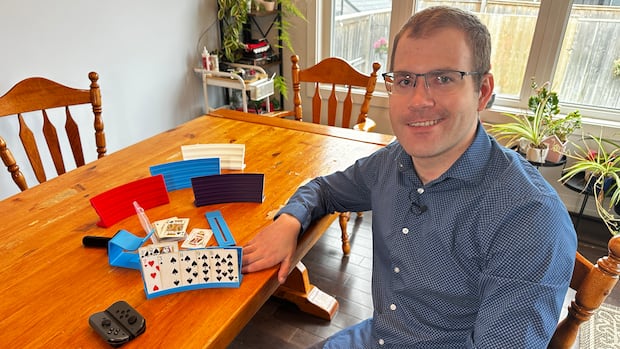 A man with glasses smiles as he sits next to a table full of 3D-printed simple tools. They include a stand to hold playing cards, an adapter to make using a nail clipper easier and an adapter that allows someone to play Nintendo Swith with one hand.