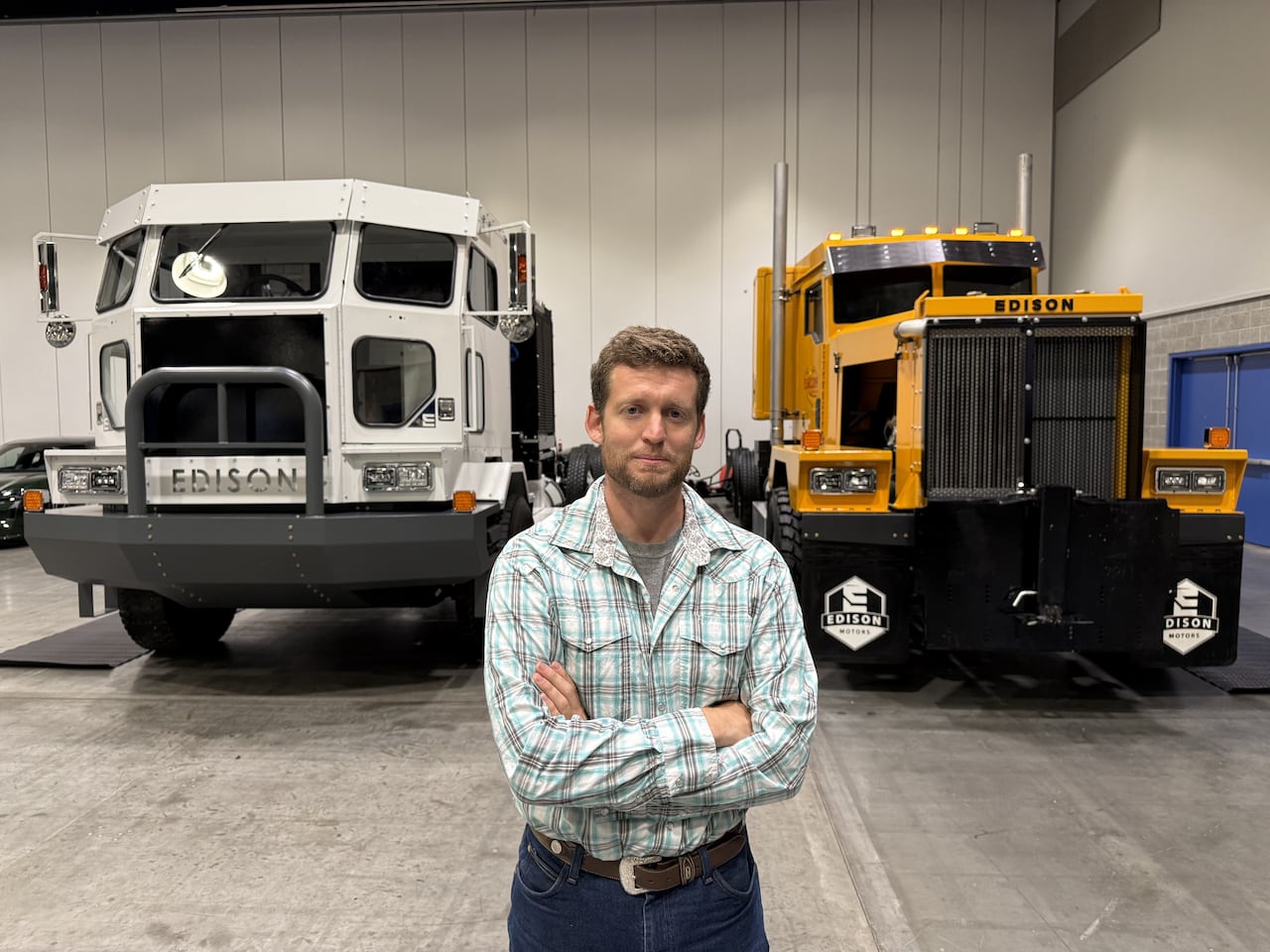 Un hombre con una camisa de cuadros azules y grises está de pie con los brazos cruzados frente a dos enormes camiones.
