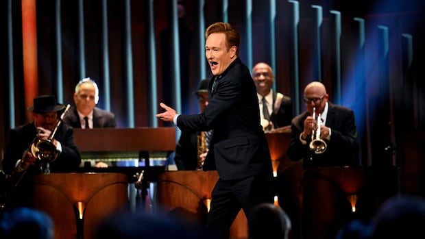 A man in a suit gestures to a band on stage.