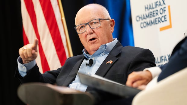 A man in a suit wearing blue glasses sits on a chair with a U.S. flag behind him.