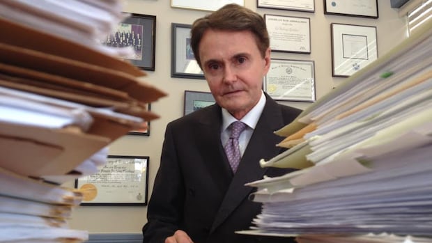 A man in a suit looks at the camera behind stacks of paper.