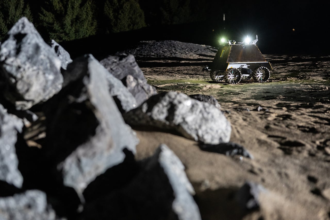 Rocks in the foreground, a rectangular robot rolling on a sandy surface in the background.