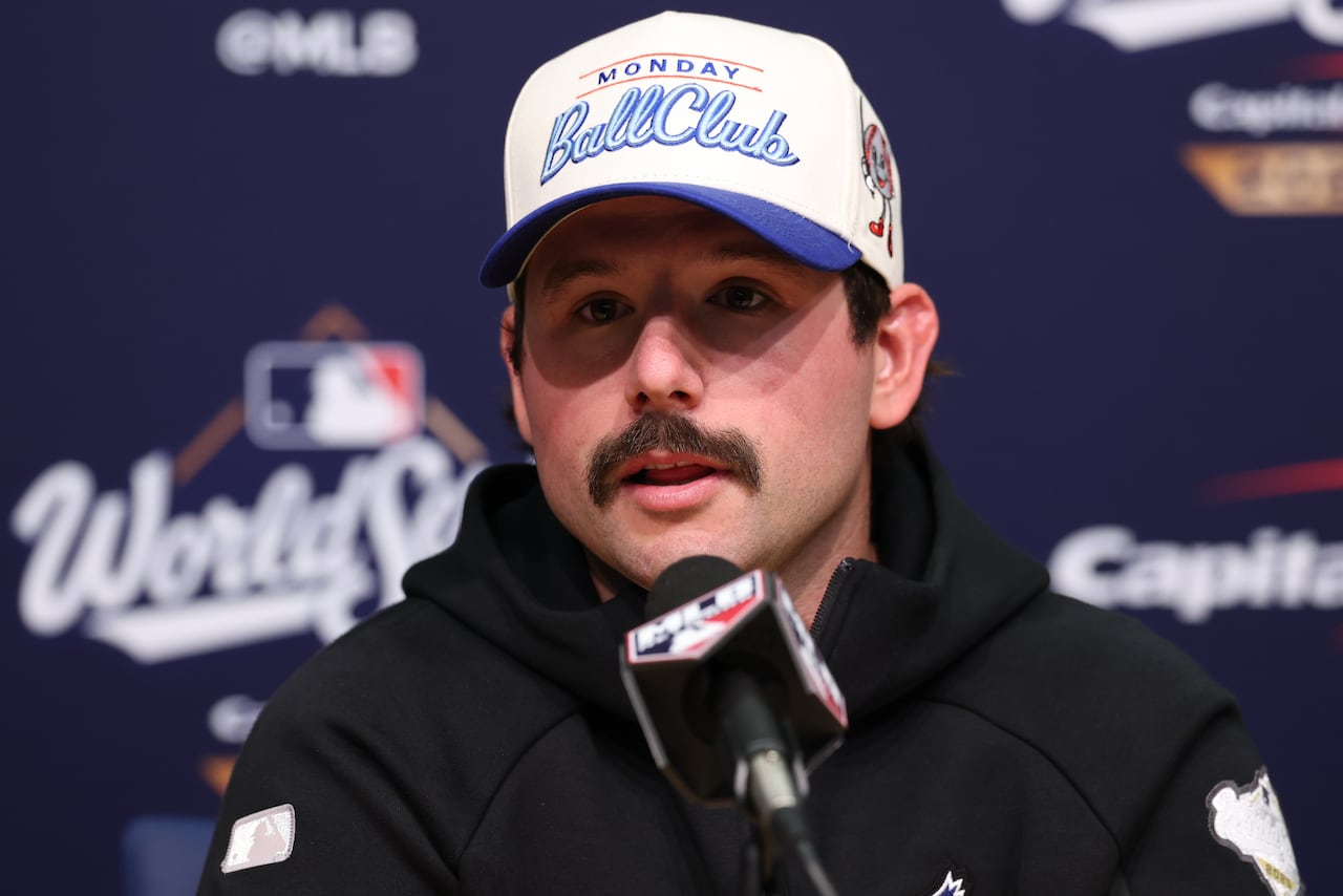 A man in a baseball cap sits behind a group of microphones.