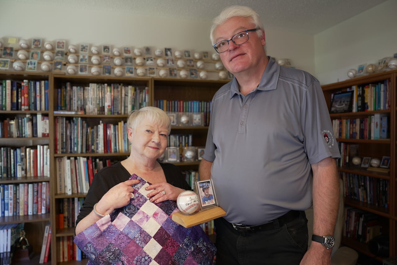 A woman holding a mostly purple patchwork quilt stands next to a man holding a baseball card and a signed baseball in a room filled with baseball books, baseballs, and signed baseball cards.