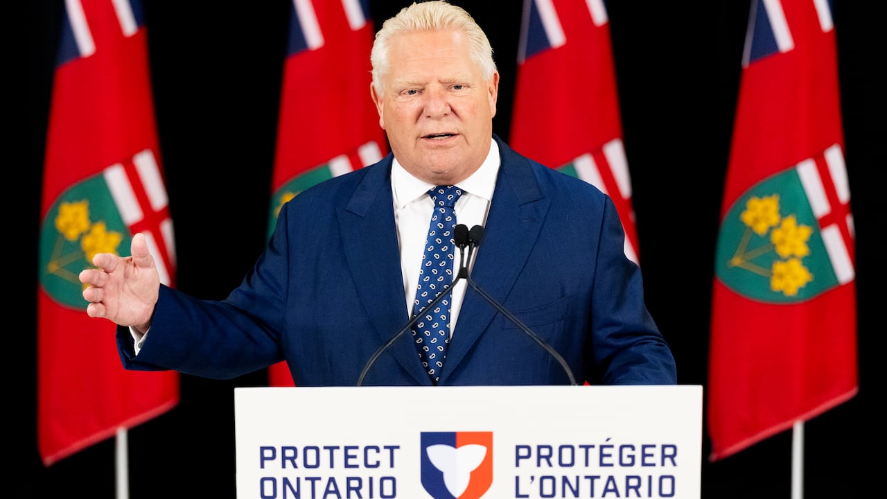 A heavyset man in suit and tie waving one hand and speaking while standing in front of a podium, with flags behind him.