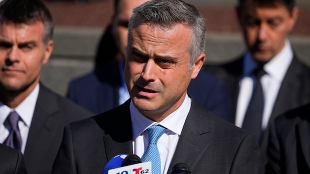 A dark haired man in a suit and tie is shown speaking in closeup in an outdoor setting. 