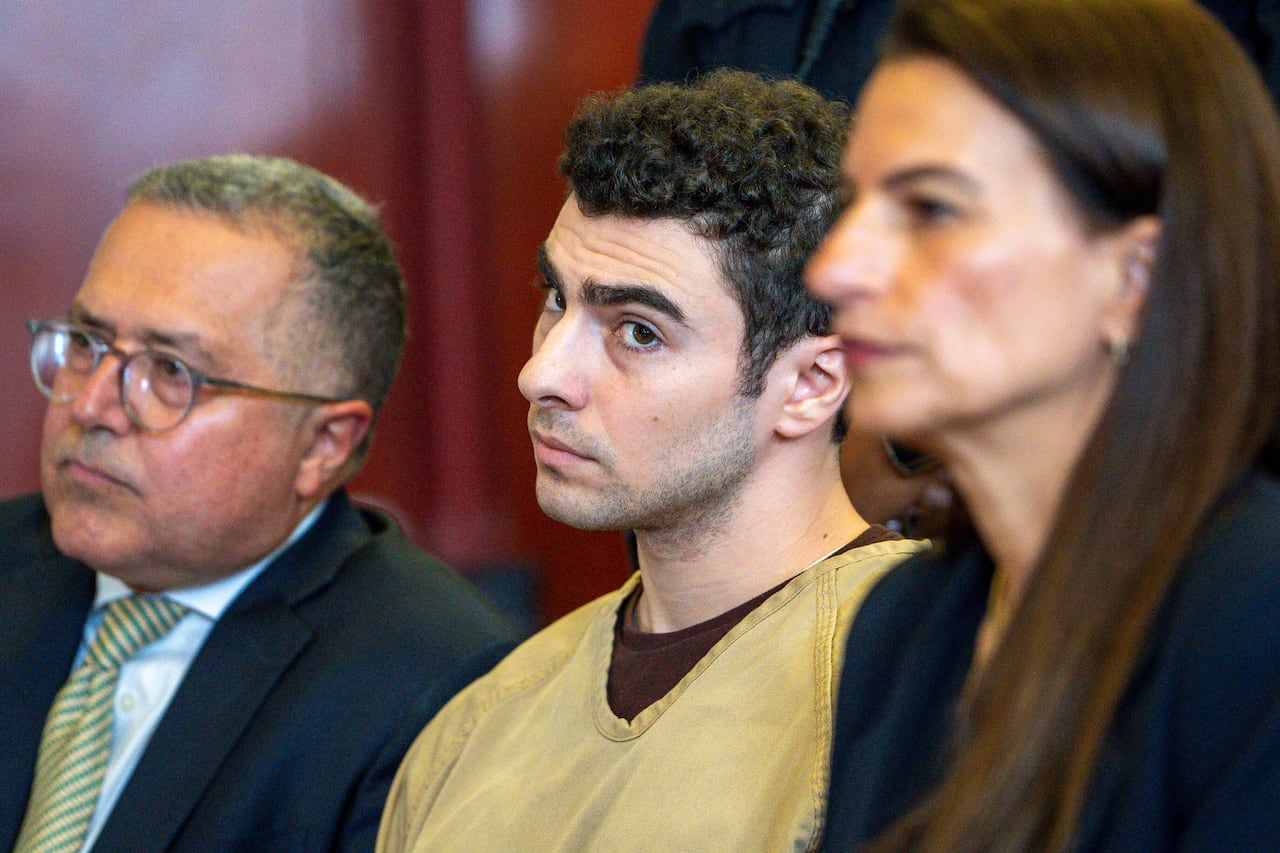 A dark haired person with a shield on his face turns his head as he sits while wearing a jumpsuit between a man and a woman.