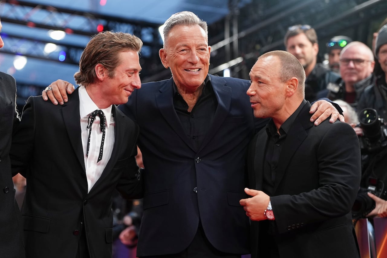 A broadly smiling man in a suit hugs two men next to him. The two men on either side appear to be talking to each other.